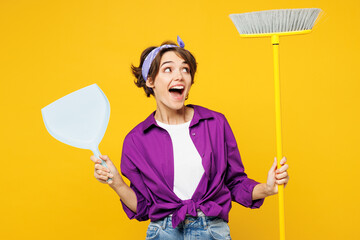 Young shocked happy fun woman wear purple shirt casual clothes do housework tidy up hold in hand brush broom scoop look aside isolated on plain yellow background studio portrait. Housekeeping concept