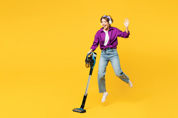 Full body young woman wear purple shirt casual clothes do housework tidy up hold vacuum cleaner listen music in headphones leap bounce isolated on plain yellow background studio. Housekeeping concept. - obrazy, fototapety, plakaty