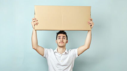 Young Adult Man in White Shirt Holding a Big Empty Cardboard Sheet Above His Head. The Model is Looking Up at the Blank Cardboard. Image with Copyspace.