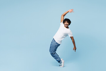 Full body young happy Indian man he wear white t-shirt casual clothes stand on toes leaning back with outstretched hands isolated on plain pastel light blue cyan background studio. Lifestyle concept.