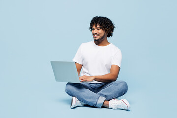 Full body young happy IT Indian man he wears white t-shirt casual clothes sits hold use work on laptop pc computer chat online isolated on plain pastel light blue cyan background. Lifestyle concept.