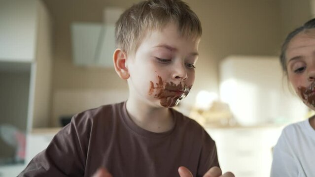 Child children eat sweet chocolate at table. Dirty face child mouth biting cocoa chocolate with teeth. Sweet breakfast of children at the table in kitchen. Happy kids with messy face eating chocolate
