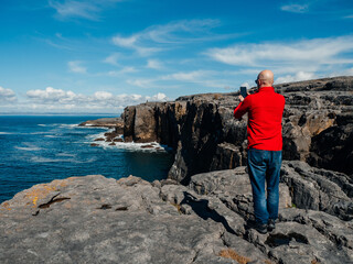 Tourist standing and enjoy view of stunning mini cliff in county Clare, Ireland. Travel and tourism. Bald man in red shirt, slim athletic body type in nature setting. - obrazy, fototapety, plakaty