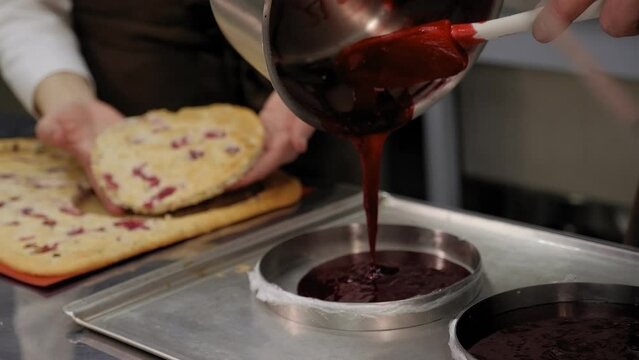 Two chefs, a man and a woman, prepare a cake in a professional kitchen, they cut a circle out of a biscuit and pour strawberry jam into a mold.