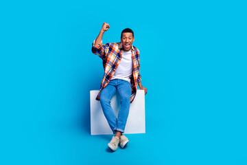 Full length photo of young man sit cube raise fist empty space wear shirt isolated on blue color background - obrazy, fototapety, plakaty