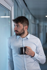 portrait d'un jeune homme employé de bureau ou homme d'affaires pensif qui fait une pause en buvant un café debout dans les couloirs de son entreprise - 775758841