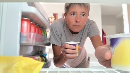 View from inside the refrigerator, adult woman, holding a jar of yogurt in one hand and taking...
