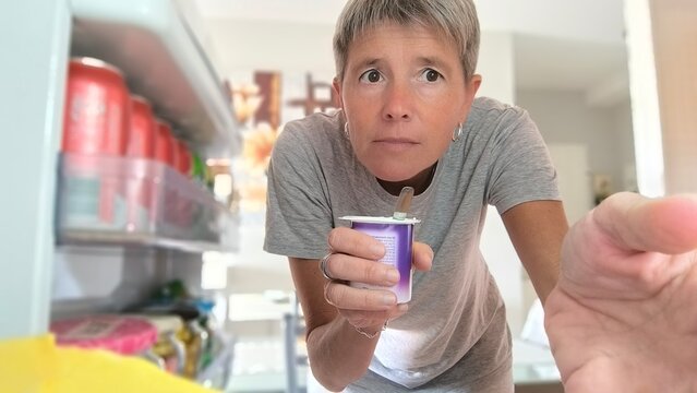 Seen from inside the refrigerator, an adult woman with short gray hair, with a determined and serious look,holding a jar of pudding with a teaspoon inside and taking something else with the other hand