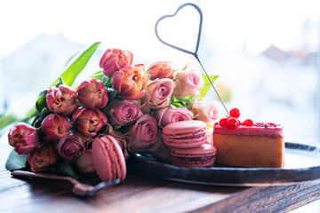 Beautiful bouquet of flowers with sweet delicacies. Heart shape, sweet pastries with pink roses and tulips on wooden table. Background for mother's day and wedding.