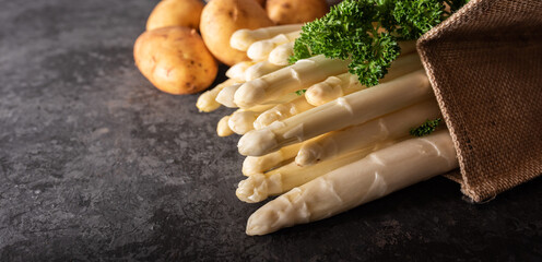 Fresh white asparagus and raw potatos in a shopping bag. Seasonal spring vegetables on black slate....