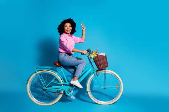Full length side profile photo of cheerful woman wear pink shirt jeans driving bicycle show v-sign isolated on blue color background