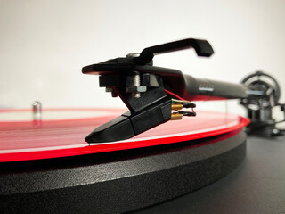 Macro closeup of a pink or red vinyl LP record on a turntable, with the tonearm needle or stylus positioned at the start of the first music track on one side of the album, ready to play.