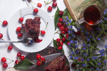 Chocolate cake with sweet cherry, cup of tea on vintage book and branch of blue flowers on white background..