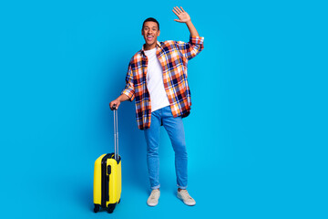 Full size portrait of nice young man suitcase arm wave hi empty space wear shirt isolated on blue...