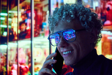Smiling curly man standing near big city mall in eyewear with urban lights reflection in glasses...