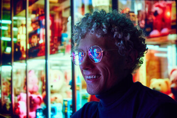 Smiling curly man standing near big city mall in eyewear with urban lights reflection in glasses. Evening city scene. Concept of emotions, lifestyle, holidays, urban life