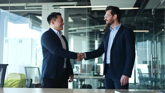 Team of two confident asian and caucasian businessmen shaking hands during meeting in well-lit office. Smiling diverse people looking at camera and gestung on background of glassy wall of cabinets.
