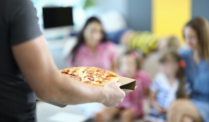 Male hands carry pizza on cardboard stand. In background, two women and two children sit on sofa.