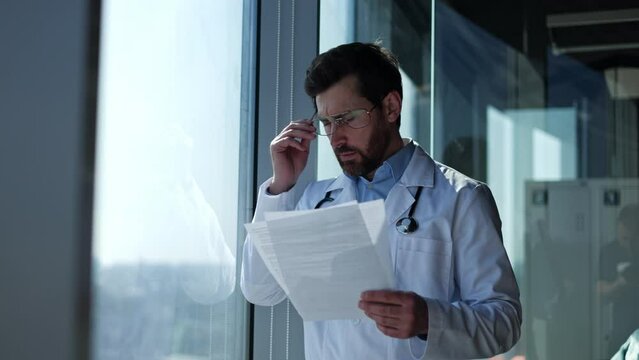 Concerned male oncologist in white coat standing by window and reading test results while getting upset. Medical worker taking off glasses while feeling disappointed because unhelpful treatment.