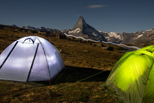 Illuminated tents glow against a moonlit backdrop by Stellisee Lake at 2600 meters, with the iconic Matterhorn mountain in Switzerland towering in the distance
