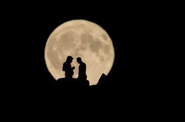 A romantic silhouette of a couple on a cliff, sharing a moment under the magnificent full moon, creating an intimate and surreal atmosphere