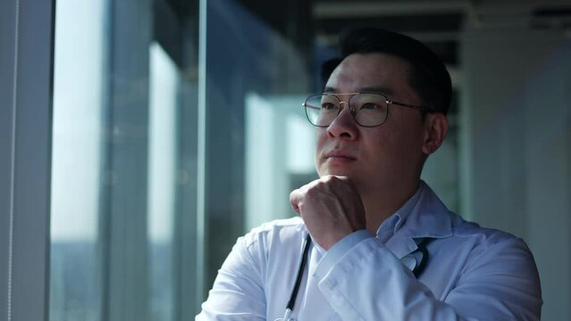 Close up of Asian male therapist in glasses touching chin with hand while standing by window lost in thought. Serious healthcare specialist pondering about optimal treatment strategy for patient.