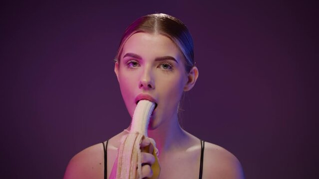 Woman takes a bite of fresh banana and licks mouth, studio shot, purple light