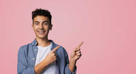 Excited happy black curly haired man in braces, wear glasses, denim shirt advertise show peek pointing area for sale slogan text, isolated rose pink background. Dental care ad advertisement concept.