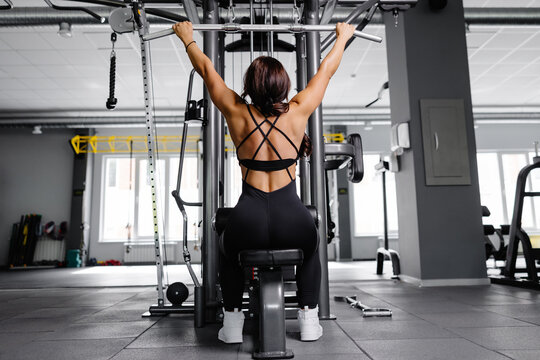Muscular athletic female bodybuilder in black suit pulled on sports simulator in gym. Back muscles are very tense.