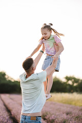 Father throws up daughter in flowers lavender enjoying scent, on summer day at sunset. Dad holds hands child girl on lavender field background. Daddy and baby embrace in meadow. Happy family outdoors.