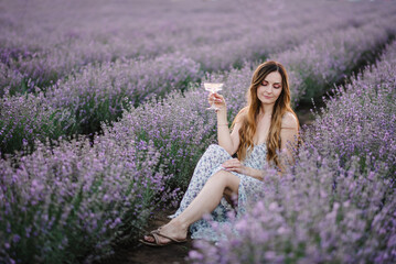 Young girl holding glass of white wine and sitting in a purple lavender field. Provence, France. Woman among violet flowers with sunlight on summer day. Female enjoying romantic time at orange sunset.