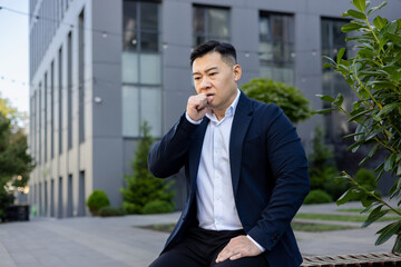 A young Asian man is not feeling well, he is sitting sick outside his office on a bench and...