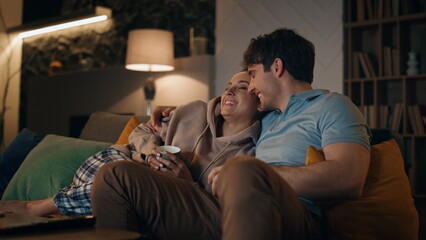 Fototapeta premium Smiling spouses watching tv sitting at cozy living room with coffee late evening
