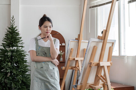 Young Asian female artist is intently painting acrylic paints on canvas in a painting studio.