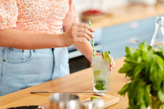 drinks and people concept - close up of woman with paper straw making lime mojito cocktail at home kitchen