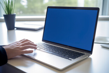 A person is using a laptop with a blue screen, placed on a white table, near a window and a potted plant - Powered by Adobe
