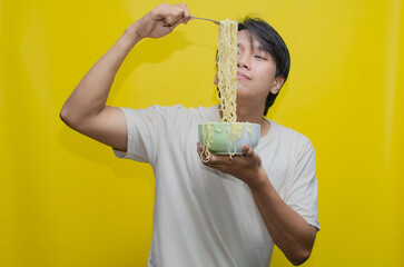 A young Asian man is inhaling and smelling the aroma of the noodles he's lifting, his expression...