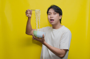 Asian Man eats noodles in a bowl by lifting them with a fork. Young Asian in cream casual cream...