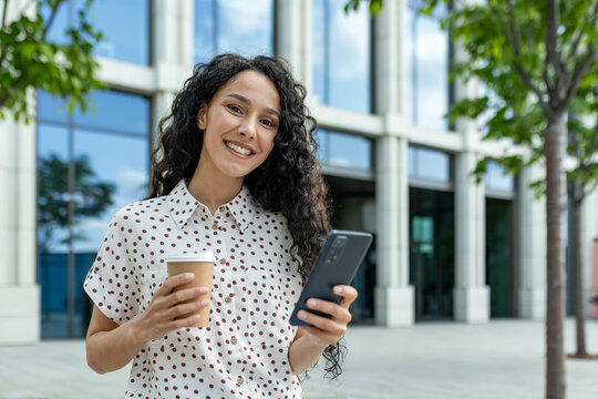 A professional woman holding a coffee cup and smartphone outside a contemporary office building on a bright day.