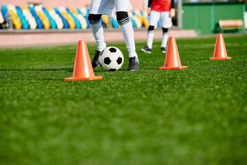 A skilled soccer player is deftly kicking a soccer ball through a series of orange cones set up in...