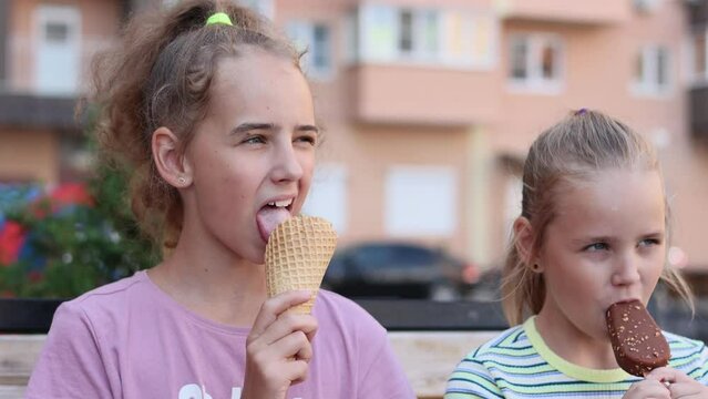 Cute little girls are eating healthy and sweet ice cream, sitting on a bench in the fresh air on a warm summer day. The ideology of summer holidays, vacations and entertainment