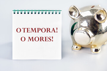 o tempora, o mores (O, the times O, the morals) Latin phrase on a white notebook on a gray background near a piggy bank in the shape of a pig - obrazy, fototapety, plakaty