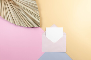 Top view of pink envelope, white card and dried palm leaf on colourful background. Blue, pink and yellow background. Colourful wallpaper, copy space.