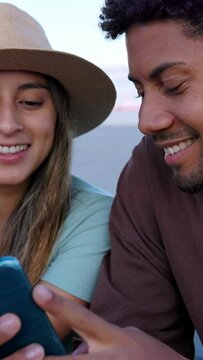 African american couple using mobile phone sitting outdoors. Two multiracial tourist friends taking city break, having fun watching social media content on app. Technology concept. Vertical video.