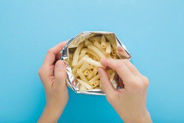 Young adult woman hand taking white sticks of potato chips from opened foil bag on light blue table...