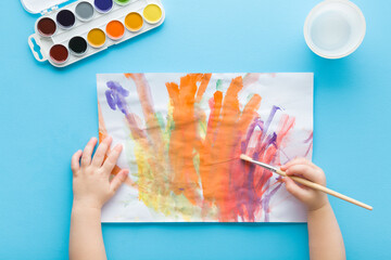 Baby boy hand holding paintbrush and drawing first colorful scratches on white paper. Pastel blue table background. Closeup. Child development. Learning painting. Point of view shot. Top down view.