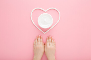 Baby girl hands and white heart shape with cream jar on light pink table background. Pastel color. Care about clean and soft child body skin. Closeup. Point of view shot. Top down view.