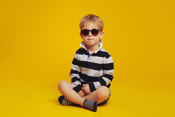 Fototapeta premium Cool little serious boy in striped shirt and trendy sunglasses feels confident while sitting crossed legged on floor against the yellow background
