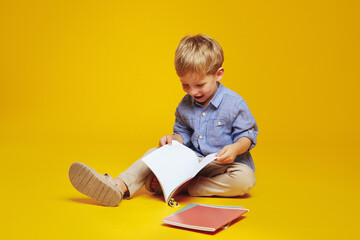 Positive curious schoolboy in casual clothes sitting on studio floor and reading notepad while...