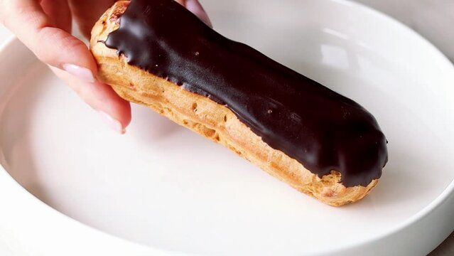 Eclair. Hand putting an eclair on white plate. Close up of woman's hand with chocolate eclair. Woman's hand putting eclair with chocolate icing on white plate, glazed dessert close-up